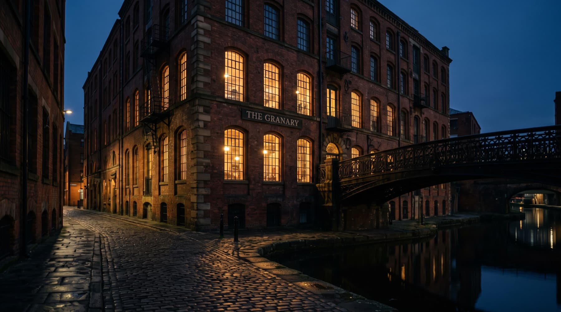 A red-brick Manchester mill beside a canal, windows glowing at nightfall